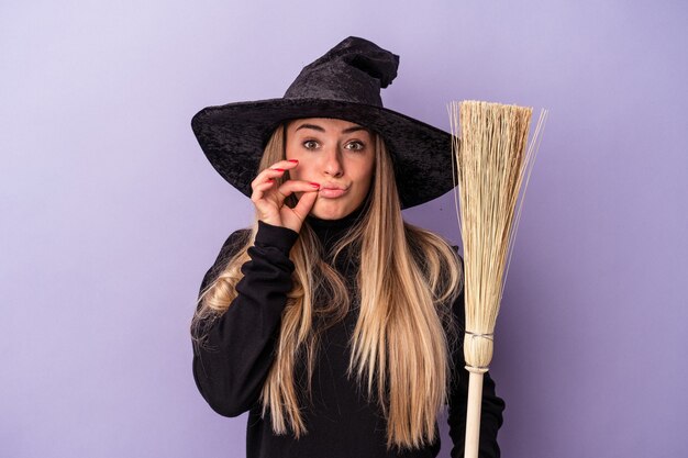 Photo jeune femme russe déguisée en sorcière tenant un balai isolé sur fond violet avec les doigts sur les lèvres gardant un secret.