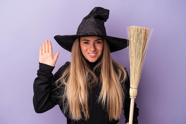 Jeune femme russe déguisée en sorcière tenant un balai isolé sur fond violet souriant joyeux montrant le numéro cinq avec les doigts.