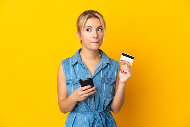 Jeune femme russe isolée sur l'achat jaune avec le mobile avec une carte de crédit en pensant