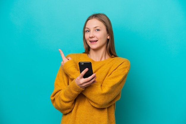 Jeune femme russe isolée sur fond bleu à l'aide d'un téléphone portable et pointant vers l'arrière