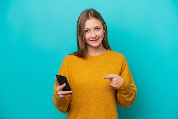 Jeune femme russe isolée sur fond bleu à l'aide d'un téléphone portable et le pointant