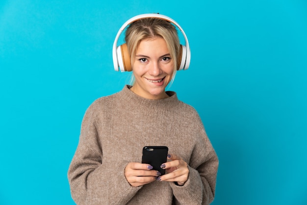 Jeune femme russe isolée sur fond bleu, écoutant de la musique avec un mobile et regardant à l'avant