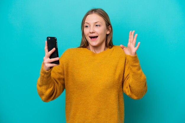 Jeune femme russe isolée sur fond bleu en regardant la caméra tout en utilisant le mobile avec une expression surprise