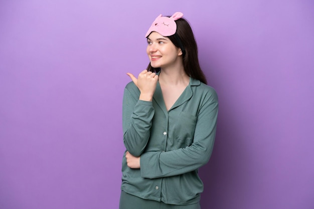 Jeune femme russe en pyjama isolée sur fond violet pointant vers le côté pour présenter un produit