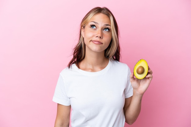 Jeune femme russe tenant un avocat isolé sur fond rose et levant les yeux