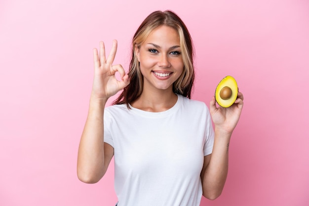 Jeune femme russe tenant un avocat isolé sur fond rose montrant un signe ok avec les doigts