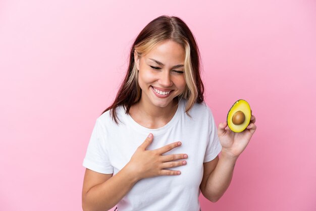 Jeune femme russe tenant un avocat isolé sur fond rose souriant beaucoup