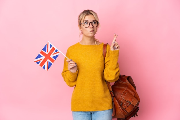 Jeune femme russe tenant un drapeau du Royaume-Uni isolé sur fond rose avec les doigts croisés et souhaitant le meilleur