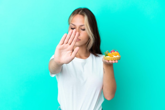 Jeune femme russe tenant un fruit sucré isolé sur fond bleu faisant un geste d'arrêt et déçu