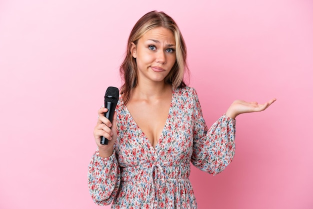 Jeune femme russe tenant un microphone isolé sur fond rose ayant des doutes en levant les mains