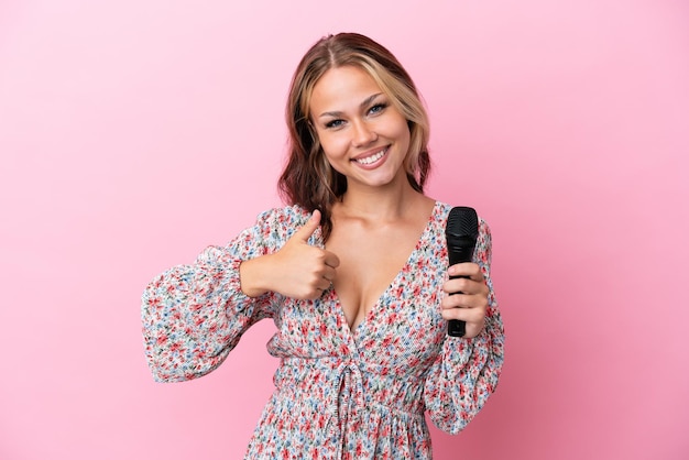 Jeune femme russe tenant un microphone isolé sur fond rose donnant un geste du pouce vers le haut