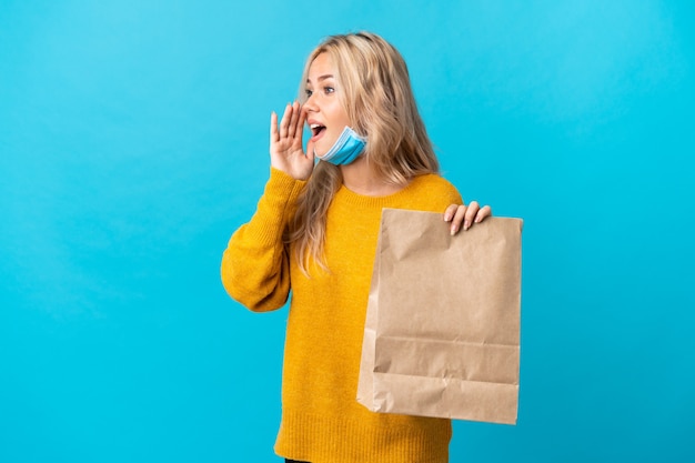 Jeune femme russe tenant un sac d'épicerie isolé sur mur bleu en criant avec la bouche grande ouverte sur le côté