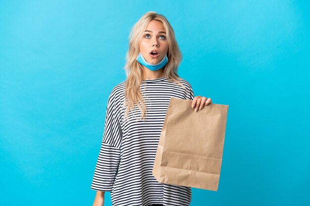 Jeune femme russe tenant un sac d'épicerie isolé sur un mur bleu à la recherche et avec une expression surprise