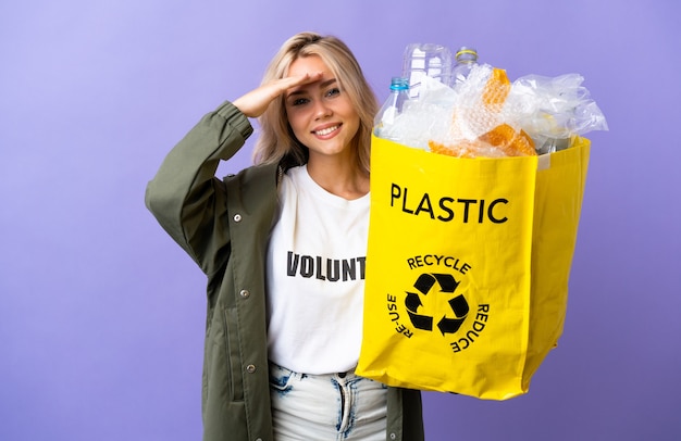 Jeune femme russe tenant un sac de recyclage plein de papier à recycler isolé sur violet à loin avec la main pour regarder quelque chose