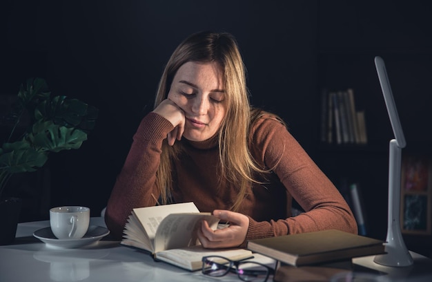 La jeune femme s'assied à la table et lit un livre la nuit avec une lumière de lampe