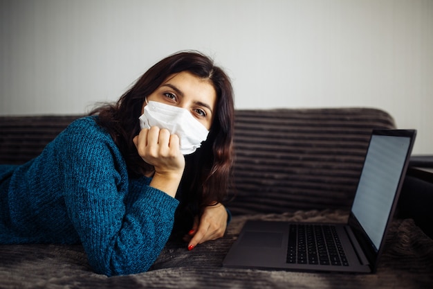 Une jeune femme s'ennuie à travailler à domicile pendant la quarantaine en raison d'une pandémie de coronavirus. Belle fille reste à la maison avec un masque médical et en tapant sur un ordinateur portable. Concept mondial de l'épidémie de Covid-19.