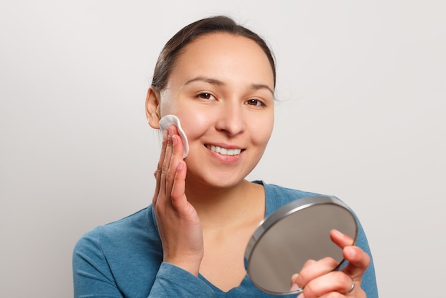 La jeune femme s'essuie le visage avec un coton avec de la lotion, regardant le miroir et souriant.