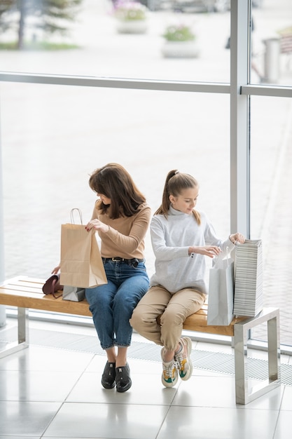 Jeune femme et sa fille assise sur un banc à l'intérieur d'un grand centre commercial et à la recherche d'achats dans leurs sacs en papier