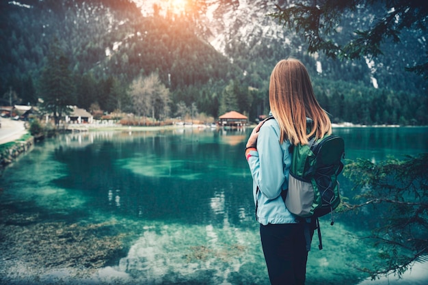 Jeune femme avec sac à dos se tient sur la côte du lac de montagne au coucher du soleil en automne. Voyage en Italie. Paysage avec fille mince, reflet dans l'eau, rochers enneigés, arbres verts en automne. Ancien