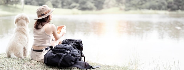 Une jeune femme avec un sac à dos voyage seule avec son animal de compagnie assis près du lac à l'extérieur