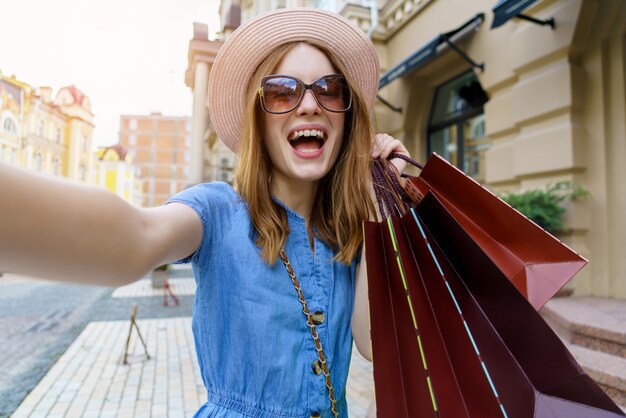 Jeune femme avec des sacs à provisions faisant selfie marchant dans une ville au jour d'été