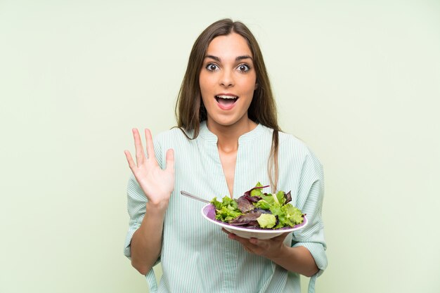 Jeune femme avec une salade sur un mur vert isolé avec une expression faciale choquée