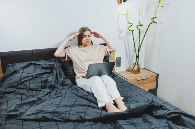 Une jeune femme satisfaite qui termine son travail sur un ordinateur portable Une fille européenne assise avec un ordinateur au lit étudie à la maison l'apprentissage à distance et le concept de travail indépendant à distance