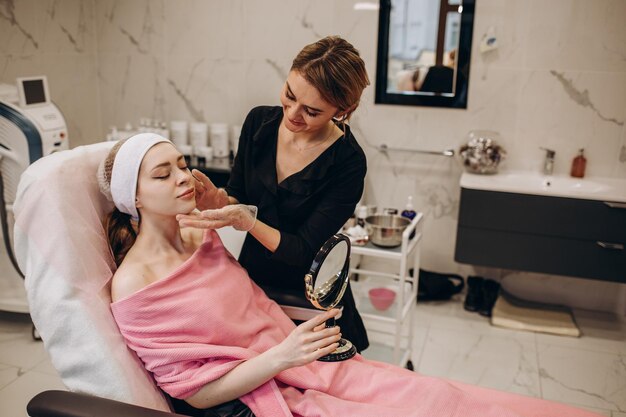 Jeune femme satisfaite regardant un miroir et touchant le visage après un traitement de cosmétologie professionnel avec une esthéticienne dans une clinique de beauté moderne