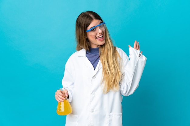 Jeune femme scientifique blonde isolée sur l'espace bleu saluant avec la main avec une expression heureuse