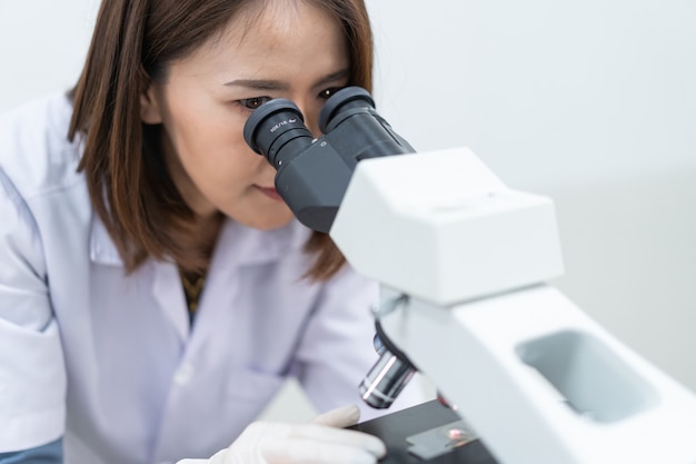 Une jeune femme scientifique en blouse de laboratoire regardant à travers un microscope dans un laboratoire pour faire des recherches et des expériences. Scientifique travaillant dans un laboratoire. Éducation stock photo