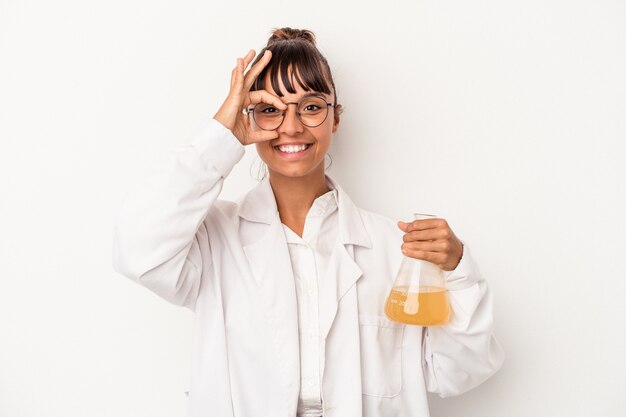 Jeune femme scientifique de race mixte tenant un tube à essai isolé sur fond blanc excité en gardant le geste ok sur les yeux.