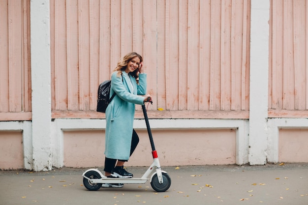 Jeune femme avec scooter électrique en manteau bleu à la ville