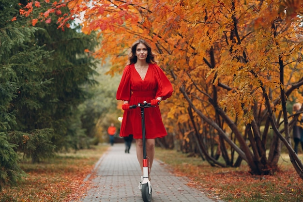 Jeune femme avec scooter électrique en robe rouge au parc de la ville d'automne.