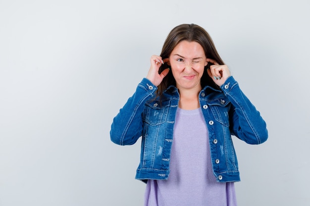 Jeune femme se branchant les oreilles avec les doigts en t-shirt, veste et ayant l'air de s'ennuyer. vue de face.