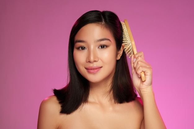 Une jeune femme se coiffe les cheveux avec un peigne de bois devant un fond rose vibrant pendant une séance de beauté