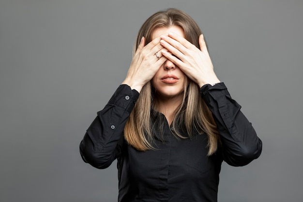 La jeune femme se couvrit les yeux avec ses mains. Belle blonde en chemise noire. Éviter la réalité et les problèmes. Fond gris.