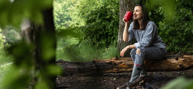 Une jeune femme se réchauffe près d'un feu éteint avec une tasse de boisson chaude dans la forêt parmi les arbres