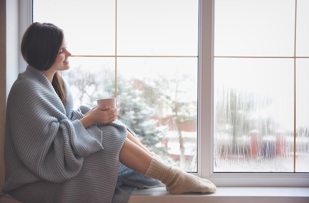 Jeune femme se relaxant en buvant du thé près de la fenêtre le jour de l'hiver