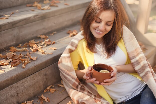 Jeune femme se reposant et buvant du thé assis dans le jardin d'automne sur les marches, enveloppé dans une couverture à carreaux en laine