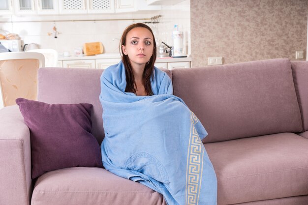 Jeune femme se sentant malade ou triste enveloppée dans une couverture bleue confortable et assise sur un canapé à la maison, regardant fixement devant elle