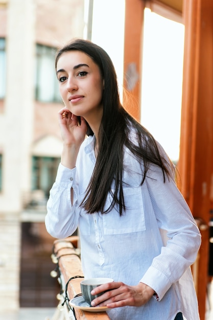 Une jeune femme se tient sur le balcon et boit du café ou du thé. La fille est calme et détendue.