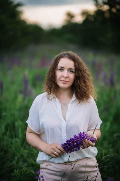 Jeune femme se tient en chemise blanche dans un champ de lupins violets et roses Belle jeune femme aux cheveux bouclés à l'extérieur sur une fleur de lupins des prés