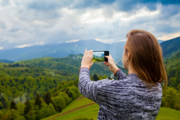 Une jeune femme se tient sur la colline et prend une photo d'un paysage magnifique avec une forêt de montagne et un ciel nuageux