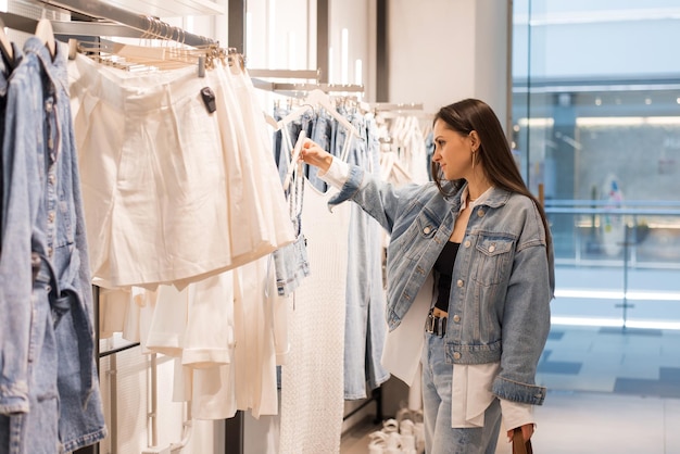 Une jeune femme se tient dans un magasin de mode et choisit soigneusement des vêtements à acheter. Elle parcourt les étagères de vêtements élégants.