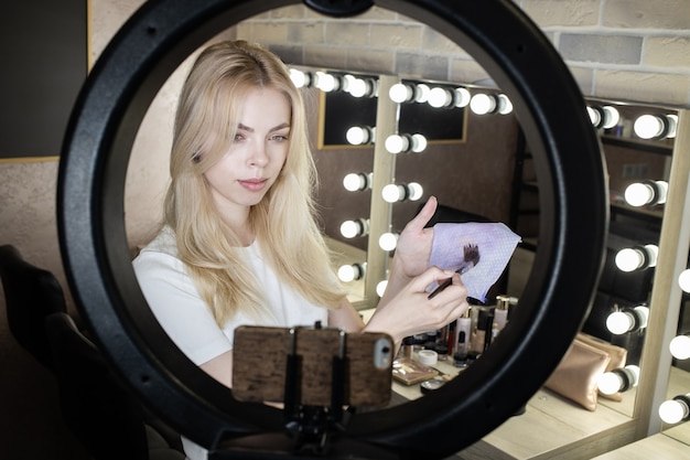 Une jeune femme se tient devant une lampe annulaire et organise des cours en ligne sur l'application du maquillage