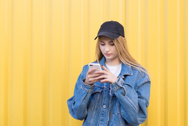 Jeune femme se tient sur un mur jaune et utilise un smartphone. écrit un message sur le smartphone et sourit.