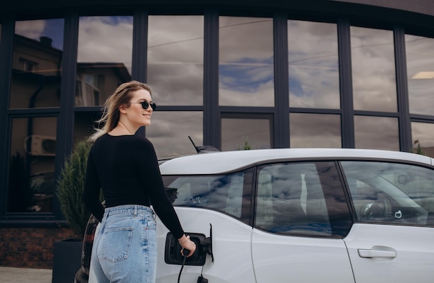 Une jeune femme se tient près de la voiture électrique et regarde le téléphone intelligent La voiture de location se recharge à la station de recharge pour véhicules électriques Partage de voiture