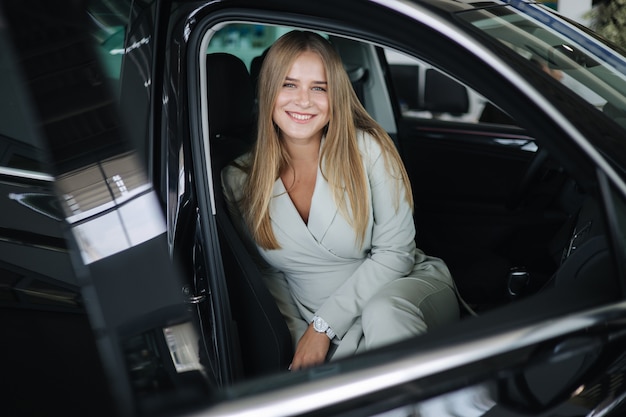 Jeune femme séduisante d'affaires assise dans une voiture dans une salle d'exposition de voitures femme choisissant une nouvelle voiture belle blonde