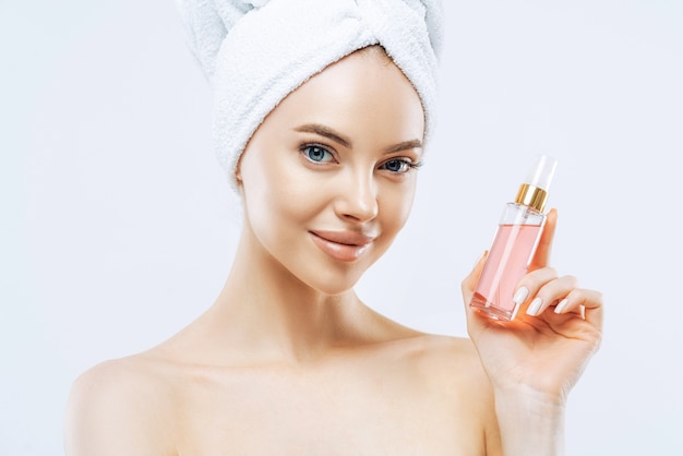 Une jeune femme séduisante applique un parfum, jouit d'un parfum agréable, se tient avec les épaules nues, a un maquillage naturel, une peau saine, une serviette enveloppée sur la tête après la douche. Excellent arôme, essayez ceci.