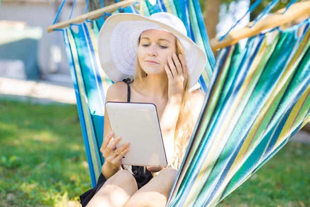 Jeune femme séduisante au chapeau à l'aide d'une tablette sur le hamac dans le jardin